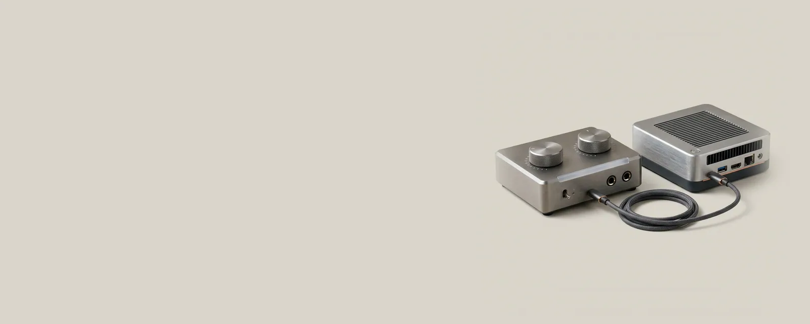Studio render of a compact aluminium audio interface with two knurled knobs, connected by a short braided USB cable to a small mini PC