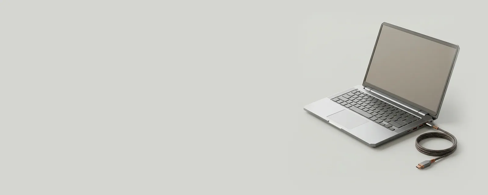 Studio render of an open generic aluminium laptop at a three-quarter angle, display showing a plain warm grey surface, a braided charging cable coiled beside it